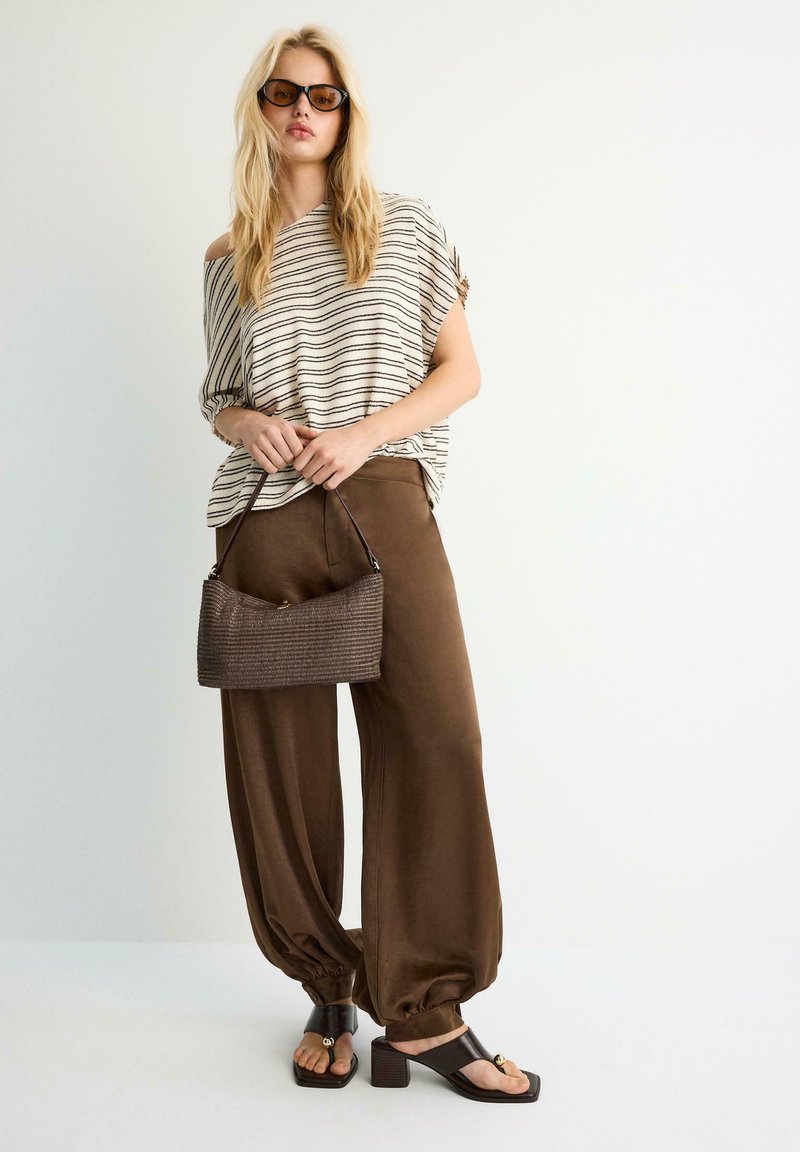 Femme blonde en haut rayé oversize, pantalons marron amples, sandales à talons noires, lunettes de soleil, tenant un petit sac à main marron texturé sur fond blanc.