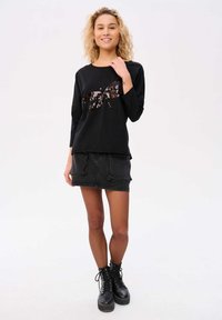 Zwarte lange mouwen top met een paillette strikontwerp, gecombineerd met een zwarte denim minirok. Model draagt zwarte veterschoenen. Eenvoudige achtergrond.