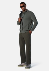 Giacca grigia con zip e due tasche frontali, abbinata a pantaloni verde oliva e sneakers grigie con dettagli blu. Il modello indossa occhiali da sole scuri.