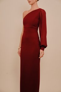 Kobieta w długiej sukni sięgającej do podłogi, z odkrytym jednym ramieniem, prawy rękaw długi i bufiaste, ubrana w złotą biżuterię na dłoniach i uchu.