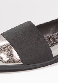 Gros plan d'une chaussure plate métallique argentée avec une large bande élastique noire à l'avant et une finition texturée et craquelée.