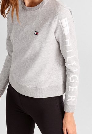 Kvinde iført lysegrå Tommy Hilfiger sweatshirt med logo på brystet og stort brandnavn på venstre ærme, kombineret med sorte bukser.