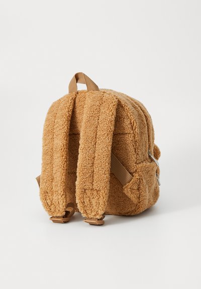 Sac à dos en polaire marron avec des bretelles rembourrées, une poignée supérieure et des poches latérales. Il comporte une fermeture zippée et un extérieur doux et texturé.