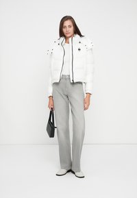 White puffer jacket with black zip and snap accents, gray straight-leg jeans, and white footwear. Model holding a small black bag.