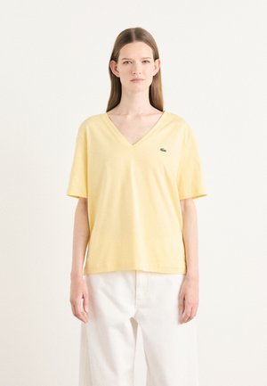 T-shirt en coton jaune avec un col en V, des manches courtes et un logo brodé vert sur la poitrine gauche. Texture lisse et coupe décontractée.