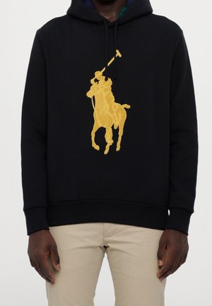 Mann trägt einen schwarzen Hoodie mit großem goldfarbenem Polo-Spieler-Logo und beigen Hosen, gezeigt von der Brustmitte bis zur Oberschenkelmitte.