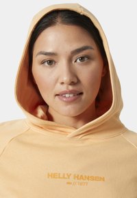 Bluza z kapturem w jasnopomarańczowym kolorze, wykonana z miękkiego materiału. Posiada haftowane logo "Helly Hansen" w nieco ciemniejszym odcieniu.