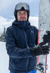 Tmavě modrá nepromokavá lyžařská bunda, černé rukavice, držící zasněžené lyže. Zasněžená krajina v pozadí, zatažená obloha se sp scattered clouds.