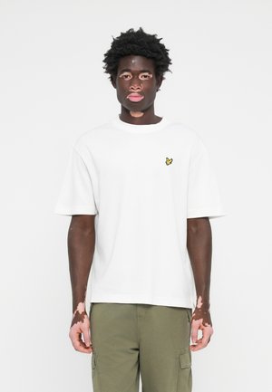 T-shirt en coton blanc à manches courtes, col rond, avec un petit emblème de papillon jaune sur la poitrine gauche. Associé à un pantalon cargo olive.