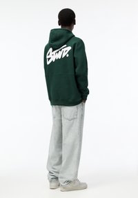 Sweatshirt à capuche vert avec un texte graphique blanc au dos, associé à un jean ample gris clair et des baskets neutres.