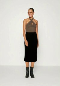 Donna che indossa un top marrone con collo ad anello, gonna midi nera, stivaletti neri e occhiali colorati, sorridente davanti a uno sfondo neutro.