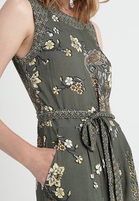 Vestido sin mangas color caqui con estampados florales, escote detallado, bolsillos laterales y un cinturón atado en la cintura con acentos geométricos.
