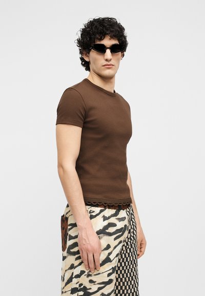 Άνδρας με σγουρά μαλλιά φοράει μαύρα γυαλιά ηλίου, στενό καφέ κοντομάνικο πουκάμισο και παντελόνι πατσγουόρκ με σχέδια ζέβρας και σκακιέρας.