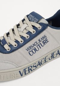 Sapatilha cinza em camurça com detalhes em pele azul-marinho, apresentando uma marcação proeminente e atacadores em bege contrastantes. Detalhes texturizados na sola.