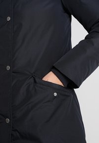 Marineblaue Pufferjacke mit Knopfverschluss und seitlichen Taschen. Das Material wirkt glatt mit einem leicht glänzenden Finish.