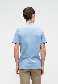 Vaaleansininen polo-paita, jossa on teksturoitu kangas, lyhyet hihat ja musta kauluksen yksityiskohta. Malli näkyy takaa, ja hänellä on päällään beiget housut.