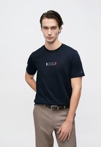 Μπλε βαμβακερό t-shirt με μικρό κόκκινο, λευκό και μπλε λογότυπο στο στήθος, συνδυασμένο με ανοιχτό καφέ παντελόνι και μαύρη ζώνη.