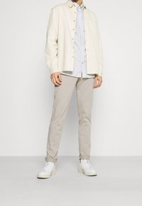 Světle béžová košile na knoflíky vrstvená přes modrou a bílou pruhovanou košili, spárovaná se světle béžovými kalhotami a bílými teniskami.