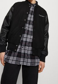 Czarna kurtka varsity ze skórzanymi rękawami, zatrzaskami, ściągaczami u mankietów. Pod spodem widoczna czarno-biała koszula flanelowa w kratę.