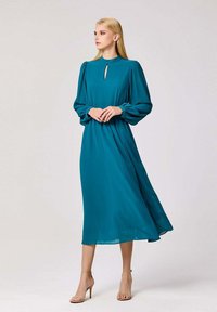 Robe midi turquoise avec un col haut, une ouverture en forme de clé, des manches bouffantes longues et un tissu fluide. Portée avec des talons nude, présentant une texture lisse.