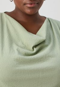 Jasnozielona, teksturowana bluzka z marszczonym dekoltem, wykonana z prążkowanej tkaniny i delikatnymi fałdami, skupiająca się na okolicy klatki piersiowej.