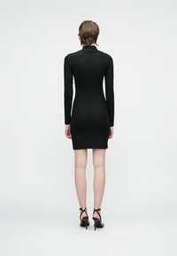 Vestido negro de manga larga ajustado con textura acanalada, cuello alto y un dobladillo por encima de la rodilla, combinado con tacones negros de tiras.