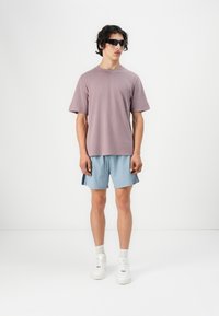 Light purple cotton t-shirt, short sleeves, paired with light blue shorts featuring dark side stripes. Model wears black sunglasses and white sneakers.