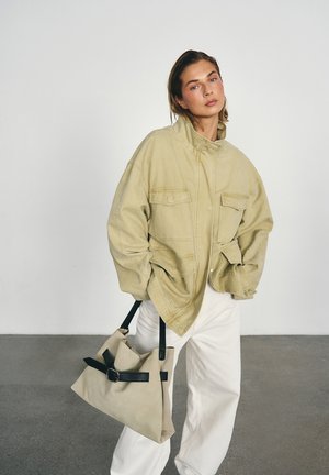 Mujer con chaqueta beige oversize y pantalones blancos, sosteniendo un gran bolso beige con asas negras, posando contra un fondo liso.
