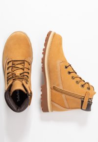 Bottines en cuir tan avec des accents marron, des lacets et une fermeture éclair sur le côté. Dotées d'une semelle en caoutchouc texturée et de coutures blanches contrastantes.