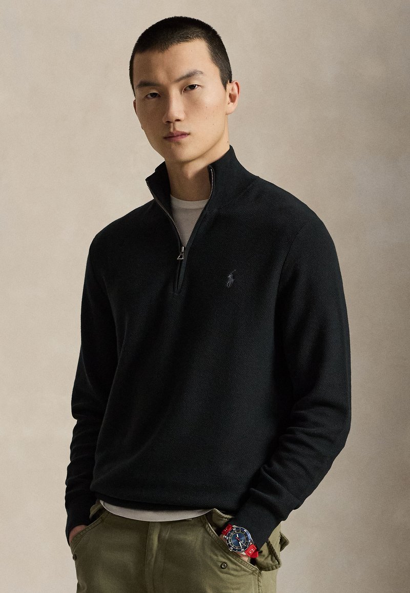Homme portant un pull noir à fermeture éclair quart, pantalon olive et une montre-bracelet bleue et rouge, debout les mains dans les poches devant un fond neutre.