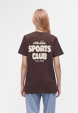 Pessoa com cabelo loiro comprido a usar uma t-shirt castanha com "ellesse SPORTS CLUB since 1959" impresso em letras brancas grandes nas costas.