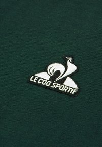 Green cotton shirt featuring a white and black embroidered logo of a bird with "LE COQ SPORTIF" text beneath. Smooth texture.