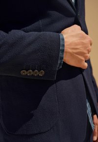 Marineblazer aus strukturiertem Stoff, vier schwarze Knöpfe am Ärmelsaum, und ein blau kariertes Hemd, das darunter sichtbar ist. Nahaufnahme von Ärmel und Hand.