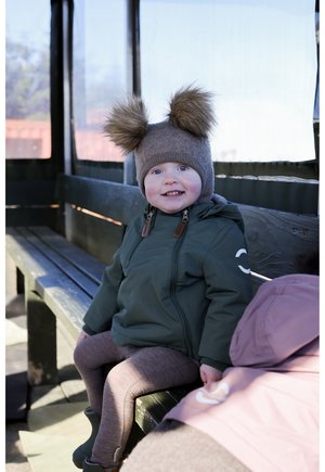 Bonnet à pompon en fourrure marron, veste imperméable verte, leggings marron. Assis sur un banc en bois, souriant à la lumière naturelle.