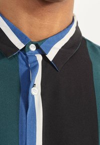 Camicia abbottonata con un motivo a righe nei colori nero, teal e blu. Presenta un colletto classico e una chiusura con bottoni bianchi al collo.