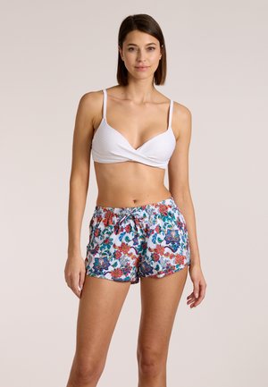 Naine seisab, kandes valget tekstuuriga bikiinitoppi ja valge lillemustrilise nööriga lühikesi pükse ühevärvilise beeži tausta ees.