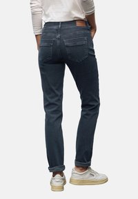 Osoba w niebieskich jeansach z mankietami i białych trampkach, stojąca z rękami w kieszeniach na jednolitym tle.