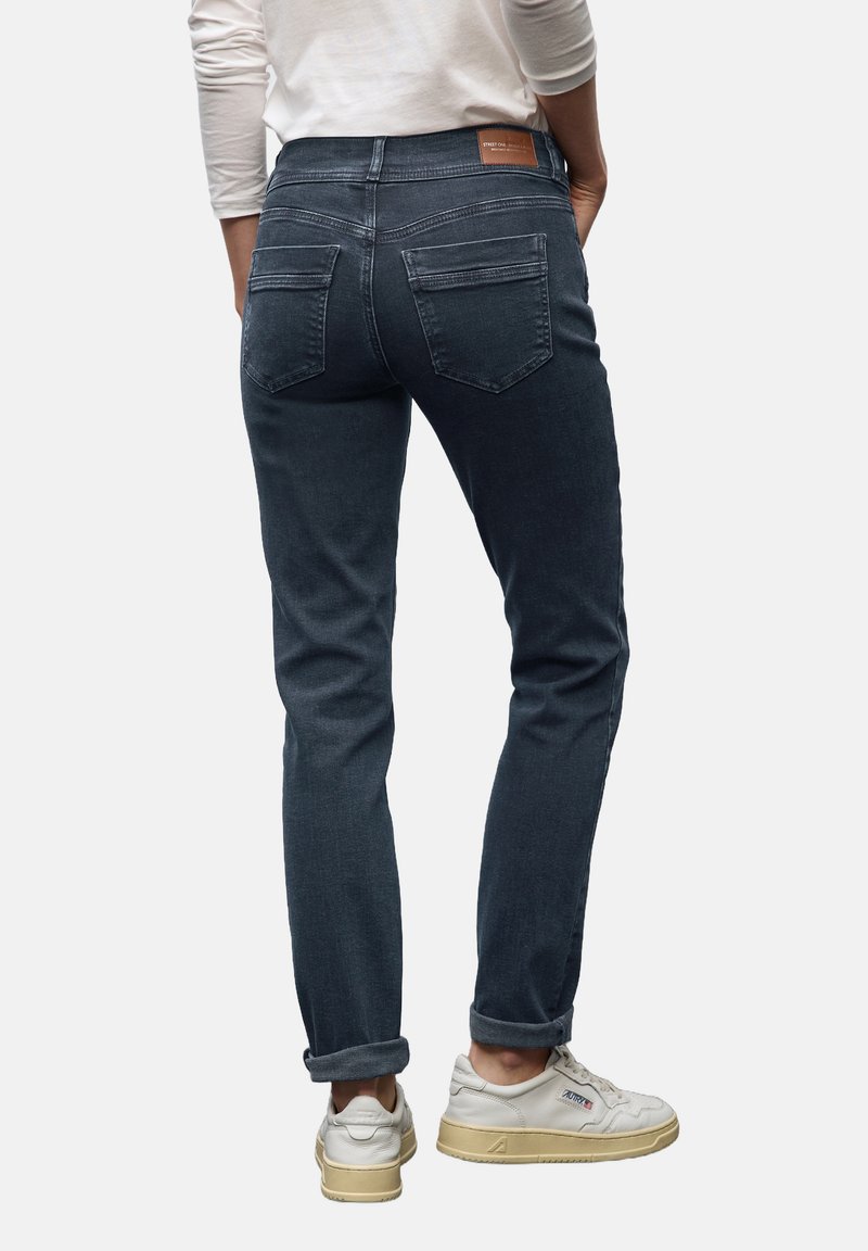 Henkilö, joka on pukeutunut käännettyihin tummansinisiin farkkuihin ja valkoisiin lenkkareihin, seisoo käsivarret taskuissa tasaisella taustalla.