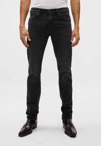 Mustad denim püksid kitsas lõikes, viie taskuga, lukuga ja kerge kulumisega; koos mustade nahast pahkluu saapatega.