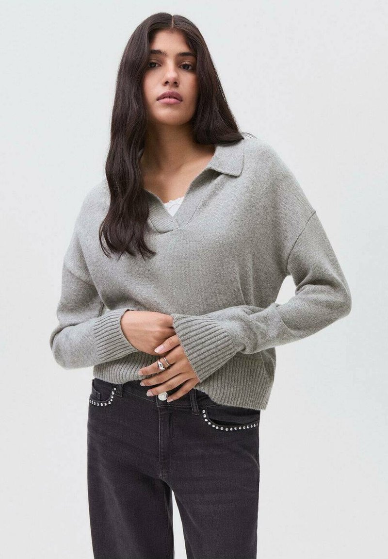 Hellgrauer, übergroßer Pullover mit kragenförmigem Ausschnitt und gerippten Bündchen. Kombiniert mit dunklen Jeans, die über silberne Nieten an den Taschen verfügen.