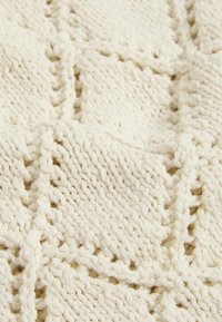 Gehaakte deken in crème kleur, met een textuur in diamantpatroon en open steken, die dikke garen en een zachte, chunky uitstraling laat zien.