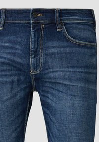 Tamno plave traperice s običnim strukom i kopčanjem na dugme, prednjim džepom i žutim šavovima.