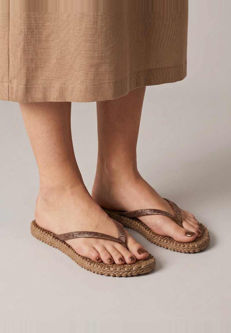 Feet wearing brown woven flip-flop sandals with metallic straps, paired with a beige skirt, on a plain light gray background.