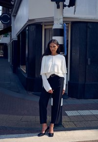 Pulover alb scurt, cu nasturi, asortat cu pantaloni negri cu talie înaltă și tocuri negre cu vârf pătrat. Fundal urban cu elemente stradale.