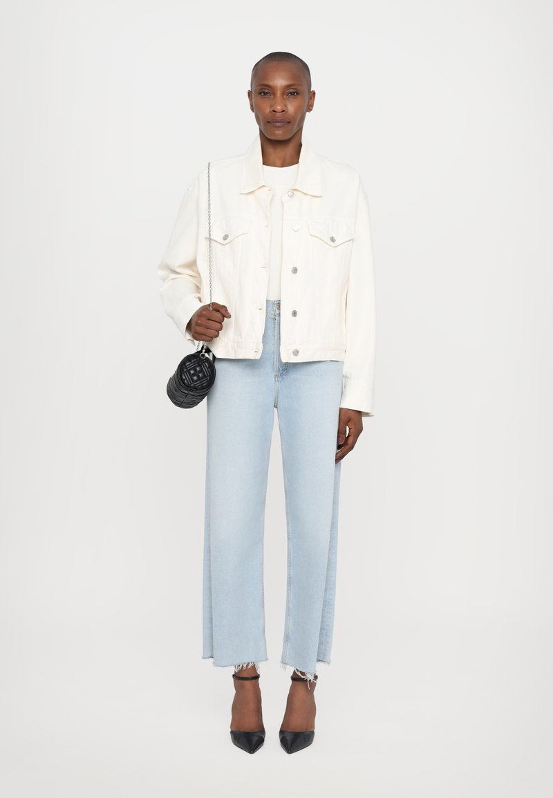 Giacca di denim bianca, jeans azzurri a gamba larga con orli sfrangiati, tacchi neri a punta e una piccola borsa nera trapuntata con tracolla a catena.