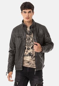 Schwarze Kunstlederjacke mit Reißverschlusstaschen, hohem Kragen und strukturiertem Finish. Tragen über einem gemusterten beigen Hemd und dunklen Hosen.