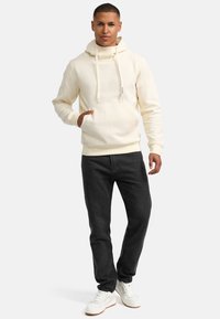Felpa a coste color crema con tasca a marsupio e cappuccio regolabile con lacci, abbinata a jeans neri e sneakers bianche.