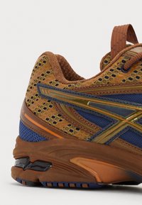 Sportskor med en brun mesh-överkropp, blå accenter och strukturerade overlay. Har en vadderad sula och en kontrasterande mörk häl-detalj.