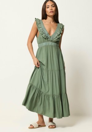 Kobieta nosząca bezrękawową oliwkowozieloną sukienkę maxi z falbankowanym głębokim dekoltem w kształcie litery V i wielowarstwową spódnicą, zestawioną z białymi sandałami wsuwanymi.