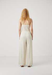Une combinaison couleur crème avec un haut ajusté présentant des détails plissés, des bretelles fines et réglables, et un pantalon large texturé s'étendant jusqu'au sol.